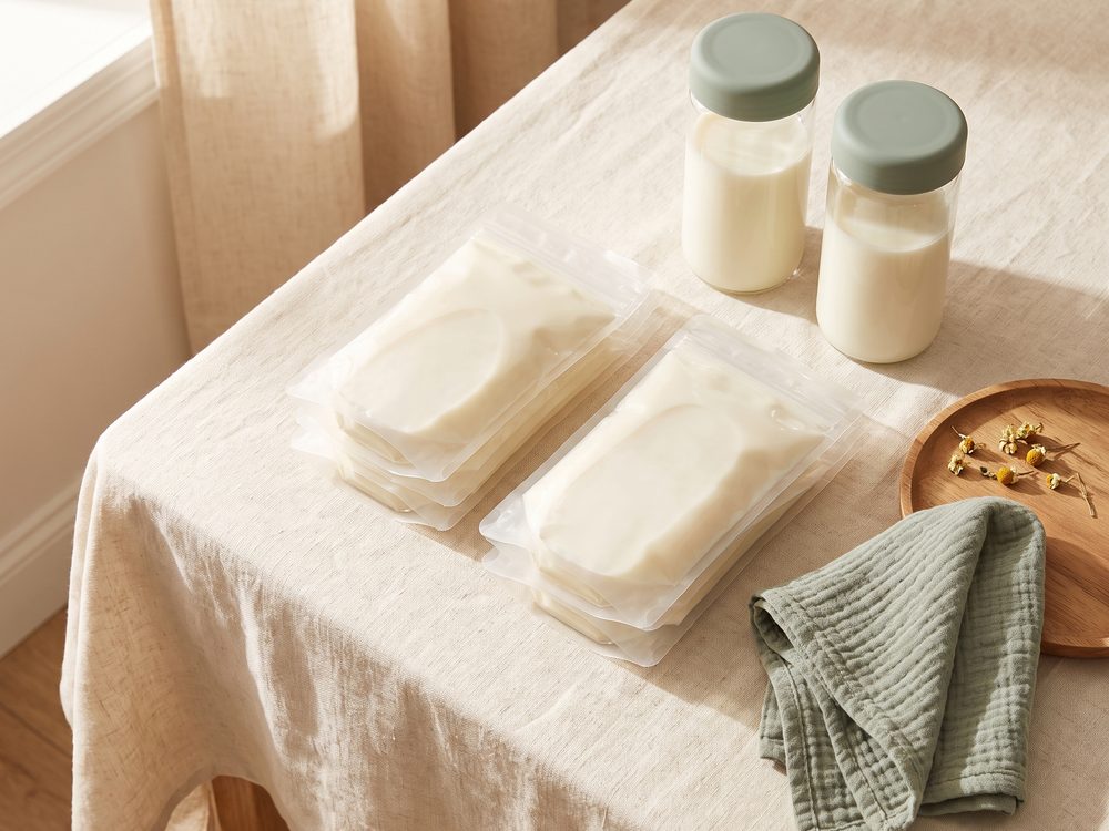 Breast milk storage bags lying flat and filled with milk beside two glass milk storage bottles with sage lids on a cream linen tablecloth in warm window light.